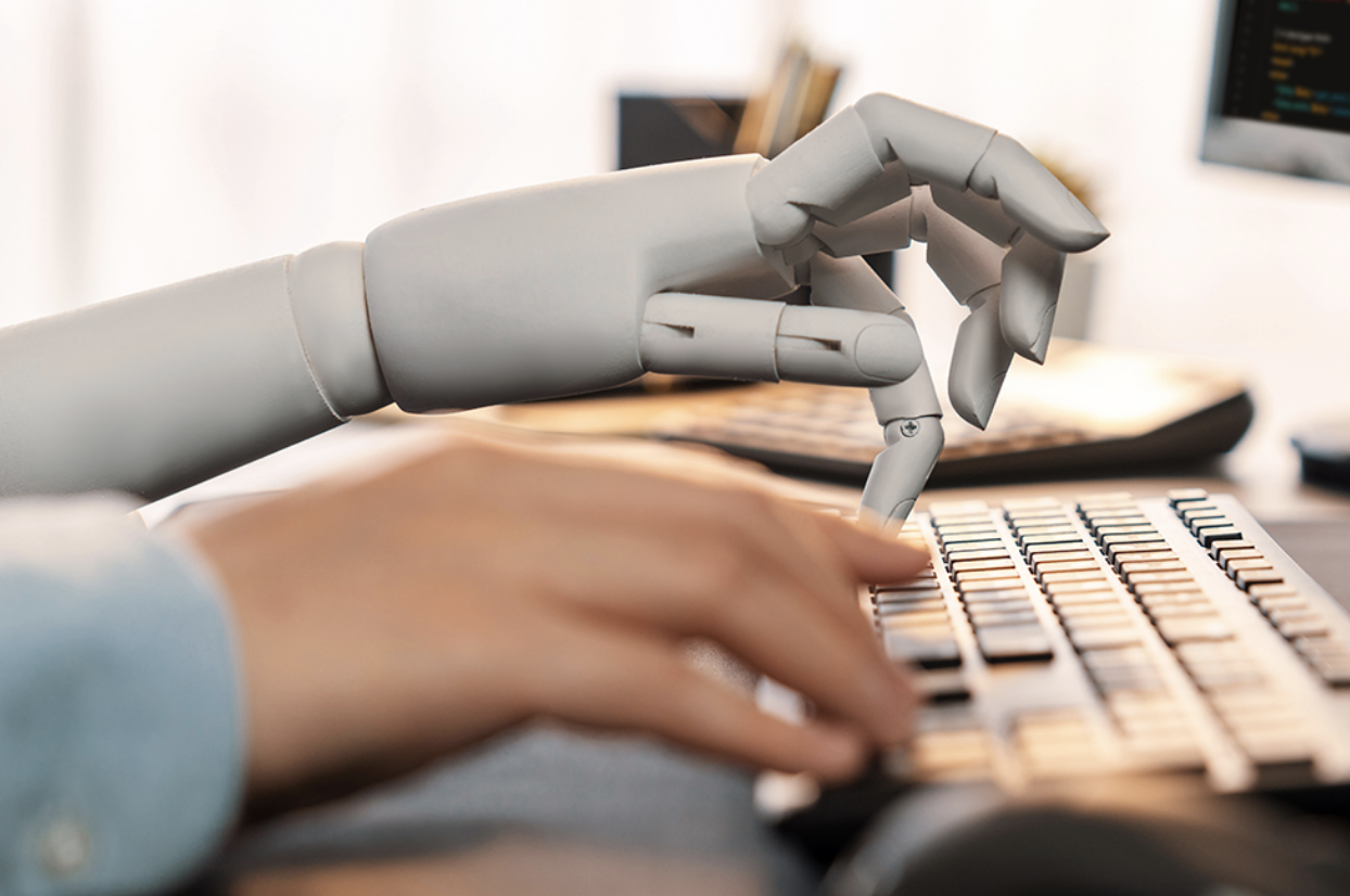 The hand of a robot next to a human hand typing on a keyboard indicating AI-assisted writing